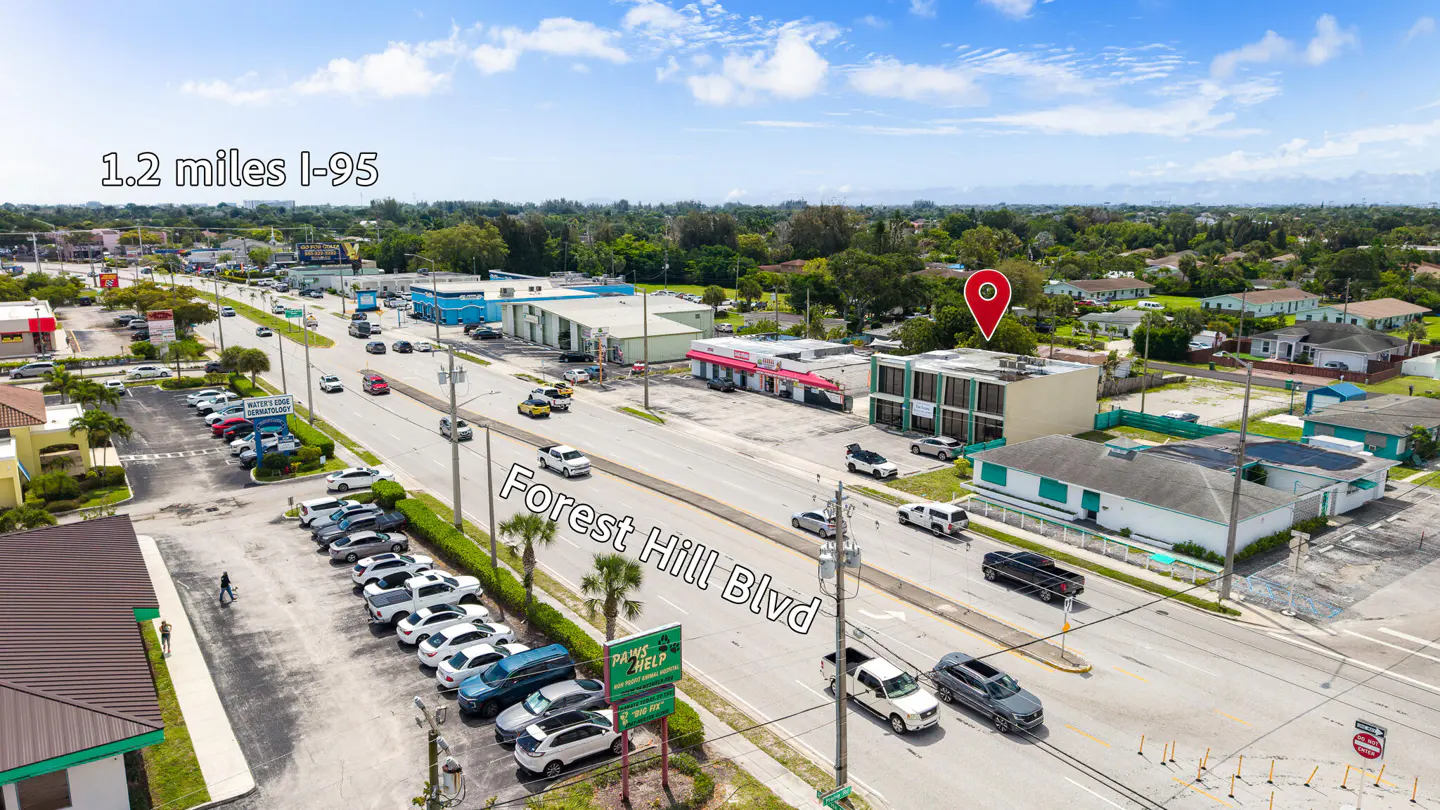 Aerial view of Forest Hill Blvd with cars, buildings, and a red location pin on a building. Blue sky with clouds.
