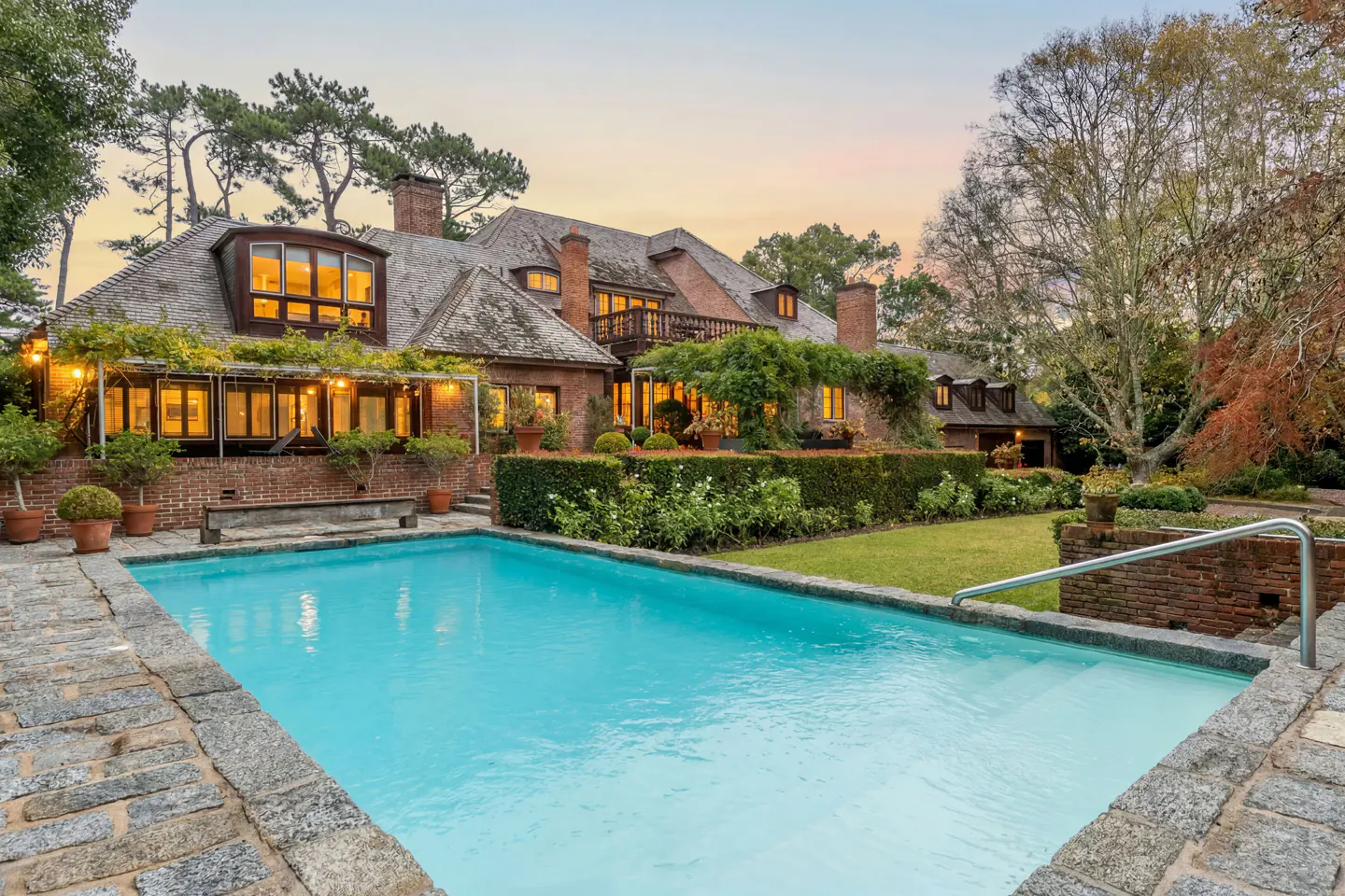 Exterior view of a brick house with a pool, green lawn, and trees at dusk.