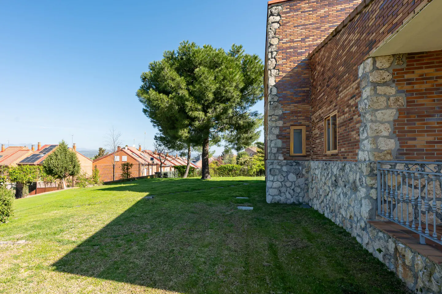 Exterior view of a brick and stone house with a green lawn, a large tree, and other houses in the background under a blue sky.
