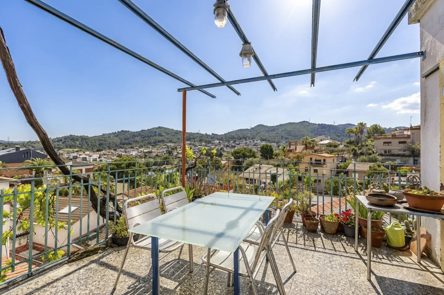 Outdoor patio with a glass table, four chairs, and potted plants overlooking a town and mountains under a blue sky.