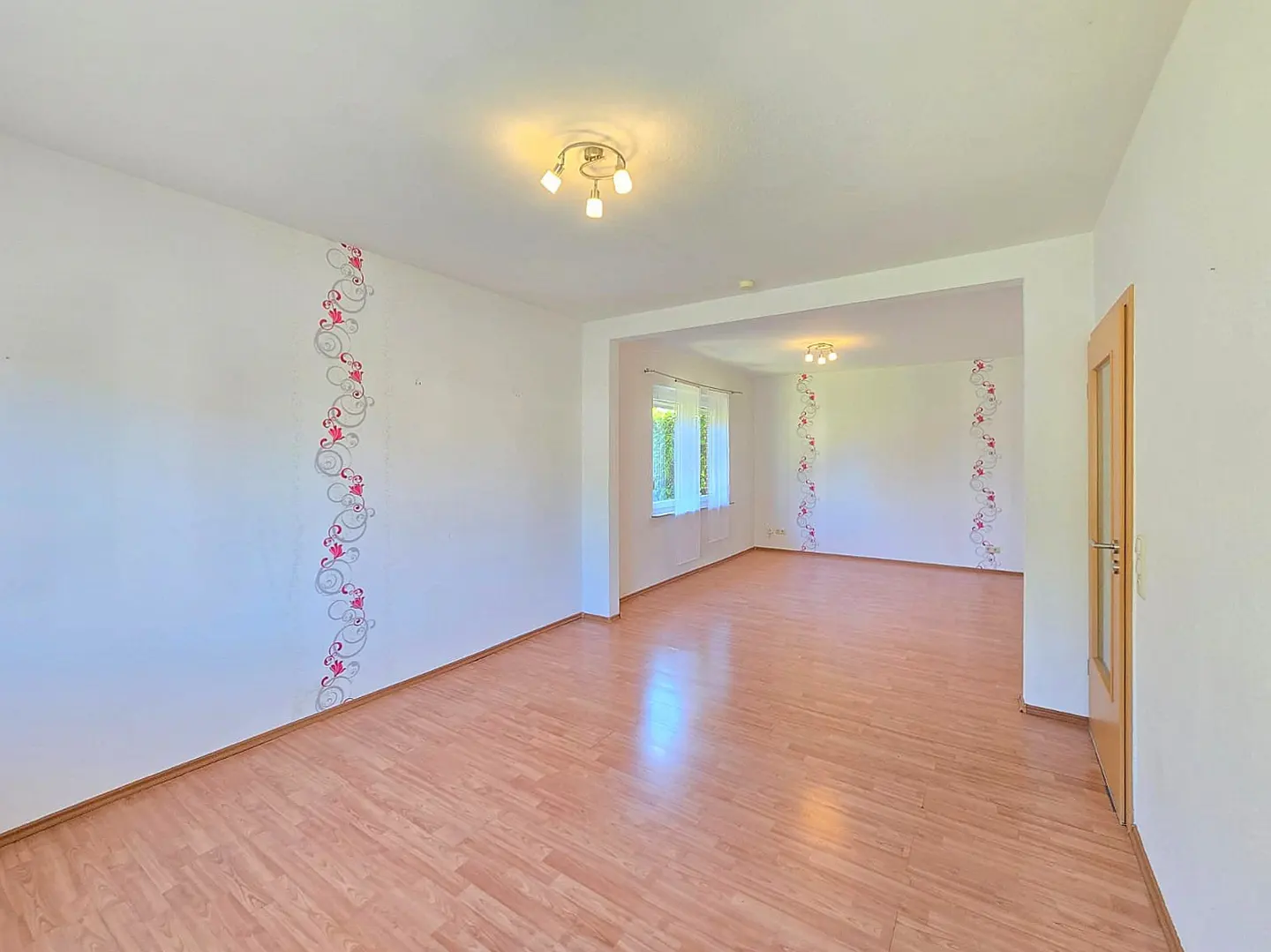Spacious, empty living room with light wood floors, white walls, floral wall decals, and modern ceiling light fixtures.