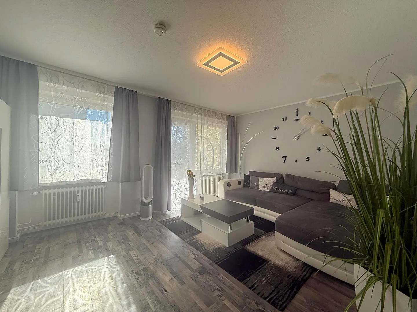 Living room with gray wood floors, gray curtains, and a brown sectional sofa. A large clock is on the wall.