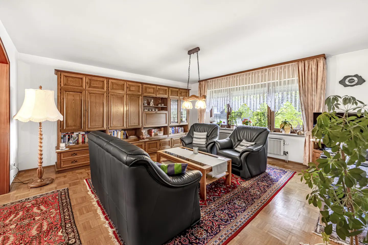 Living room with black leather sofa and armchairs, wooden wall unit, patterned rug, and a large window with beige curtains.