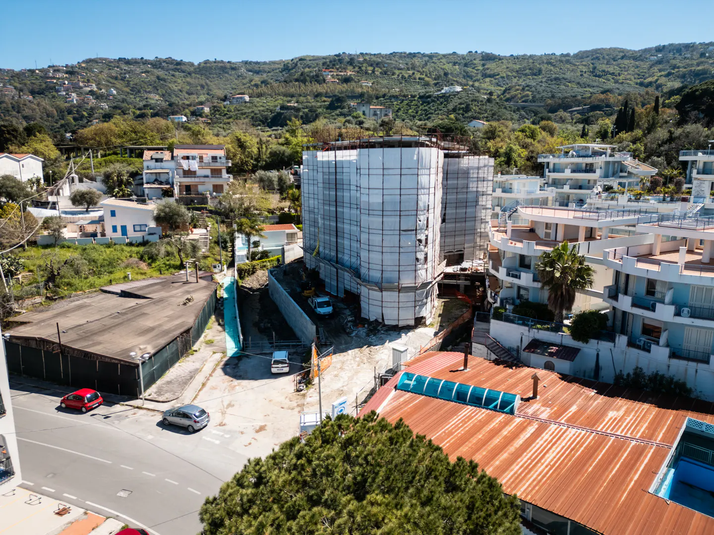 Aerial view of a building under construction, covered in white scaffolding, surrounded by houses and green hills.