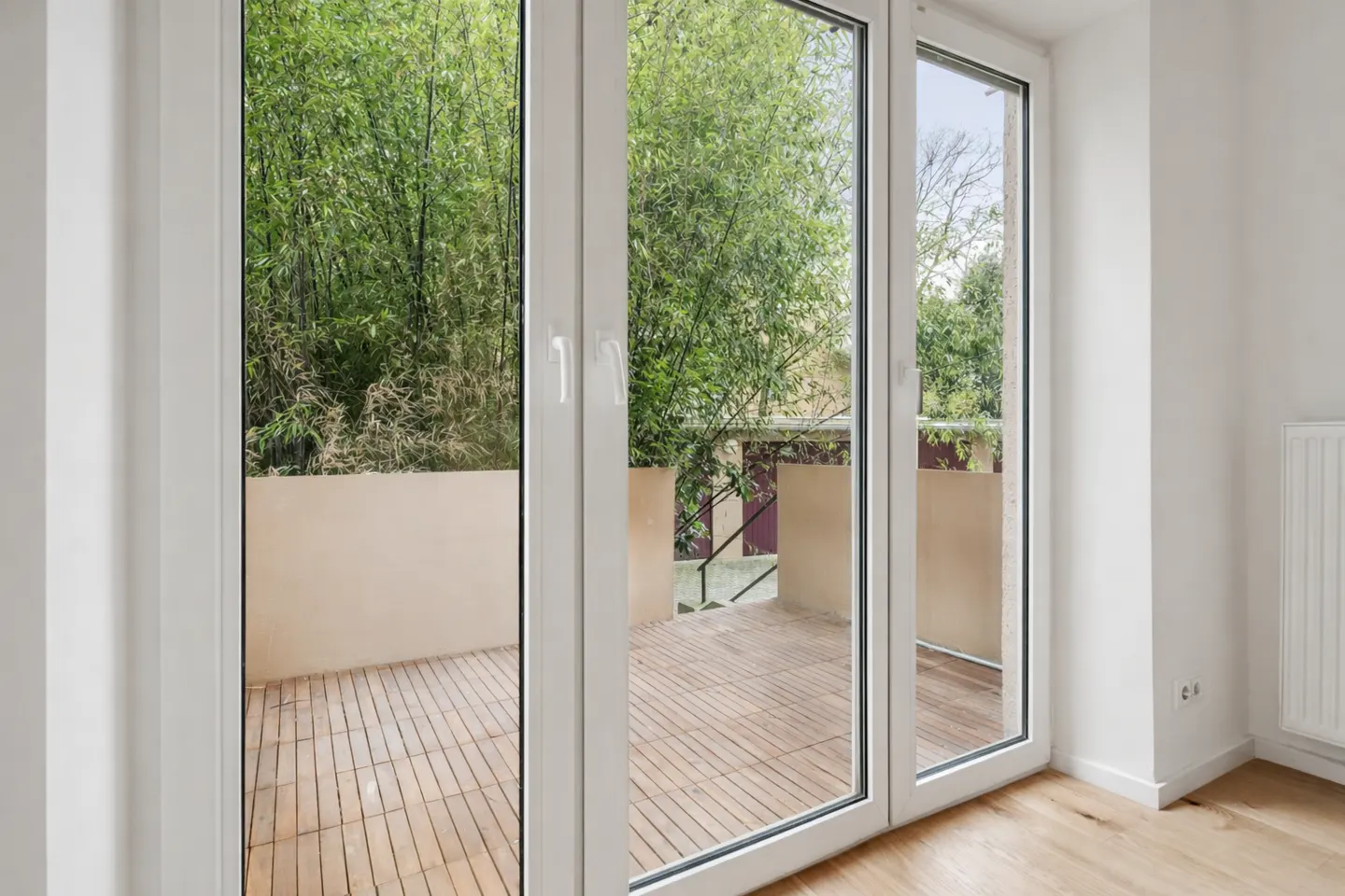 White glass doors opening onto a wooden balcony with a view of lush green bamboo trees and a beige privacy wall.