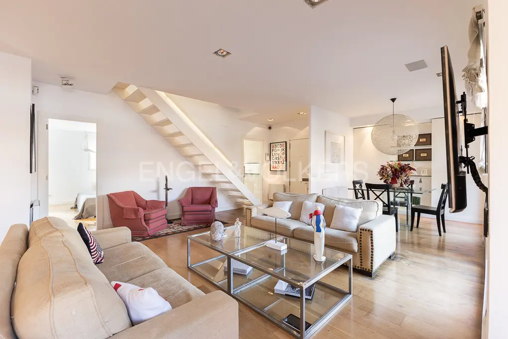 Bright living room with beige sofas, glass coffee table, and red armchairs. White staircase leads to the upper floor. Dining area visible in the background.