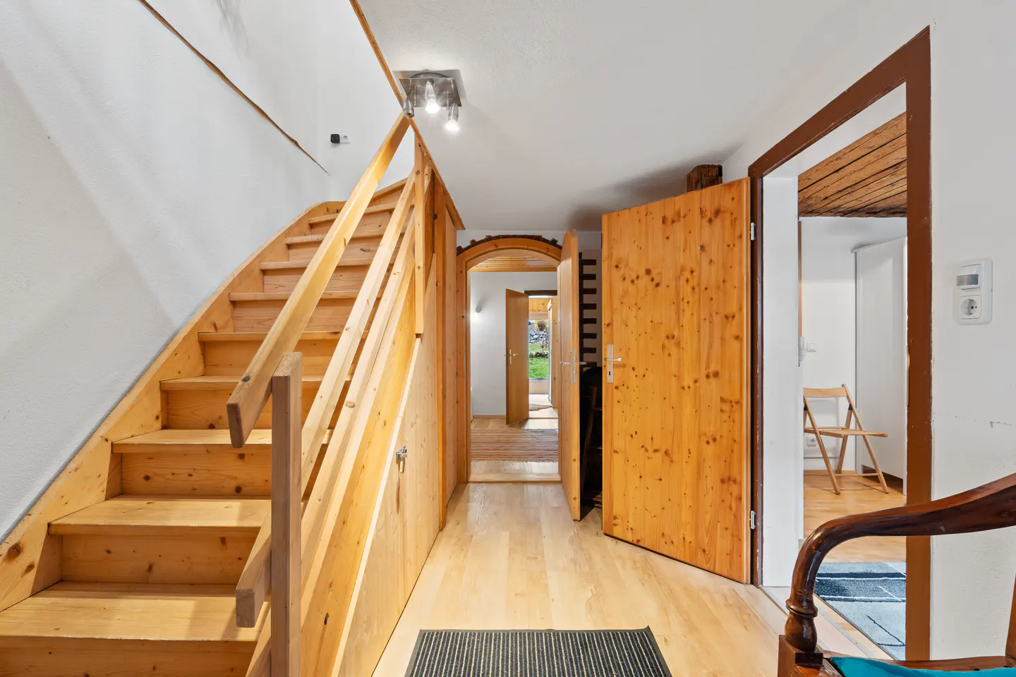 Interior view of a hallway with light wood floors, stairs, and doors. A rug is on the floor. A chair is partially visible in the foreground.