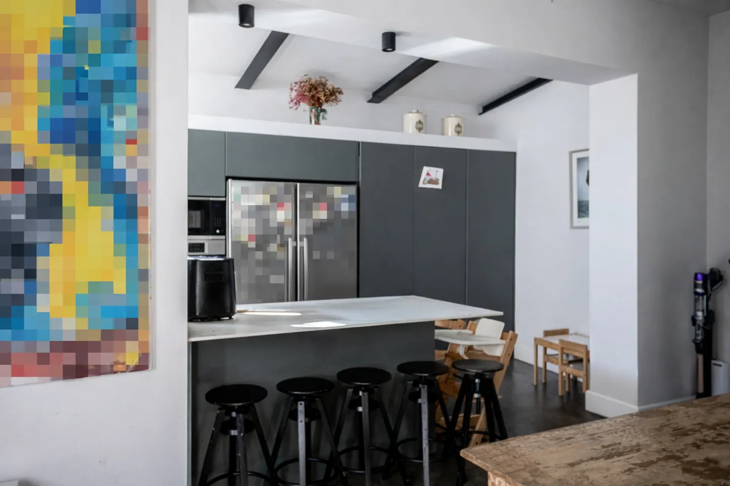A modern kitchen with gray cabinets, a white countertop island with black stools, and a colorful pixelated painting.