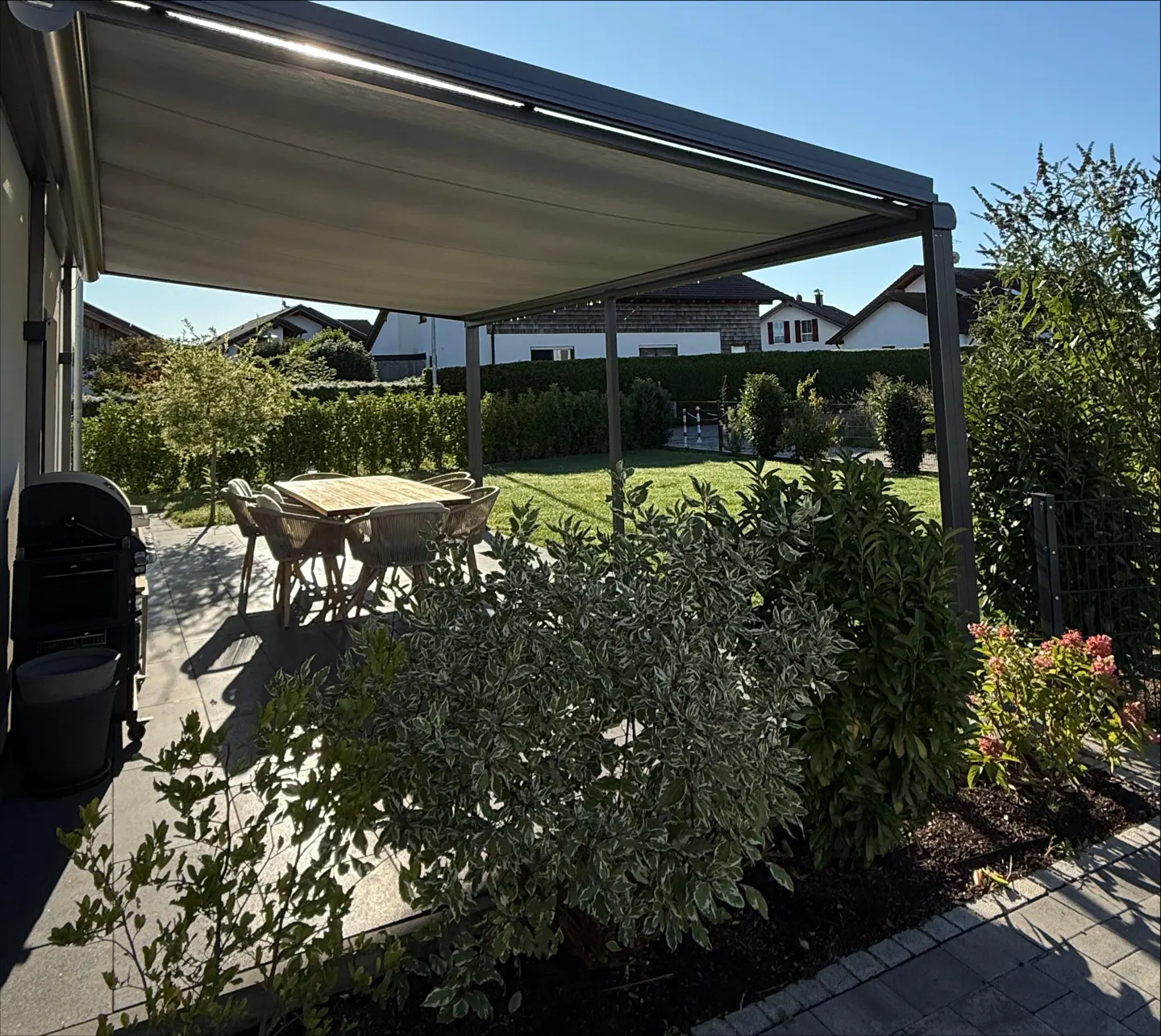 Outdoor patio with a gray pergola, table, chairs, grill, and lush green landscaping.