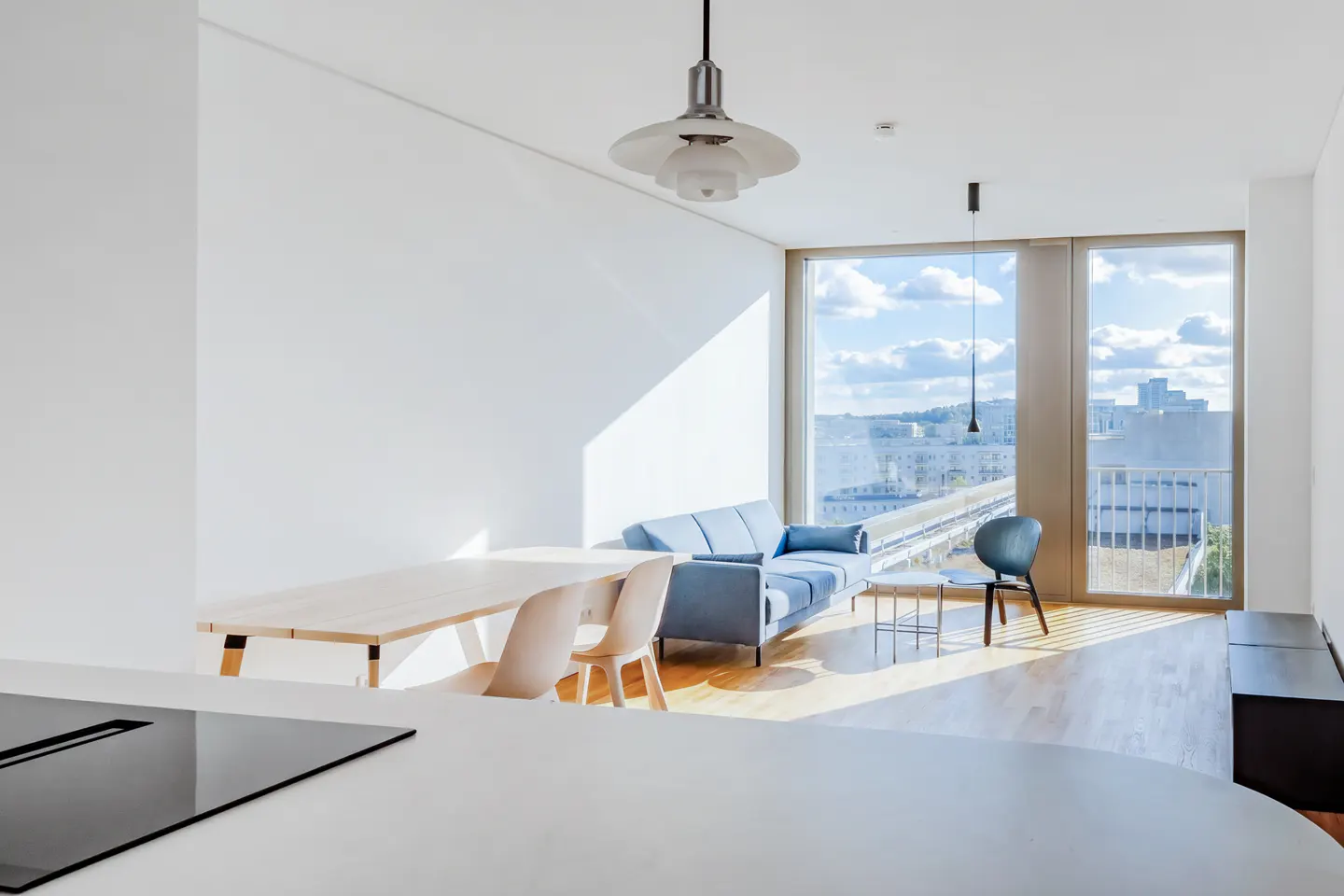 Bright, modern apartment interior with a blue sofa, wooden table and chairs, and a large window overlooking a city.
