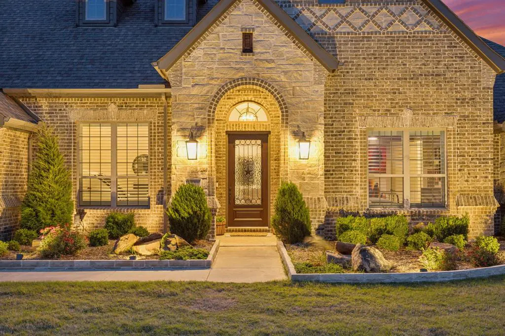 Front view of a tan brick house with a dark roof, a brown door with a star, and green bushes.