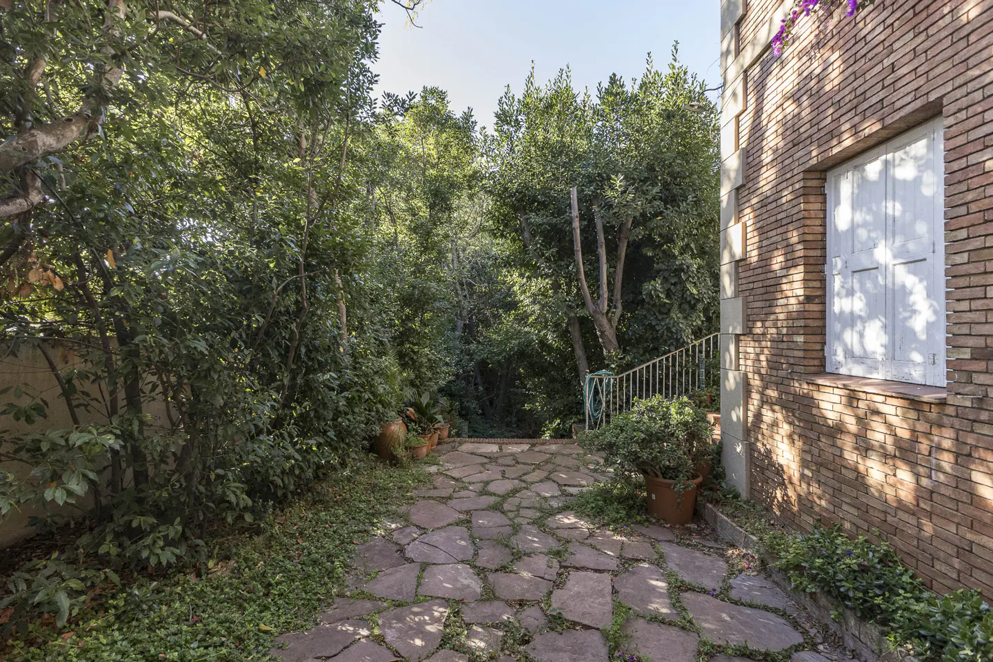 Stone path leading to a lush green garden next to a brick building with a white shuttered window.