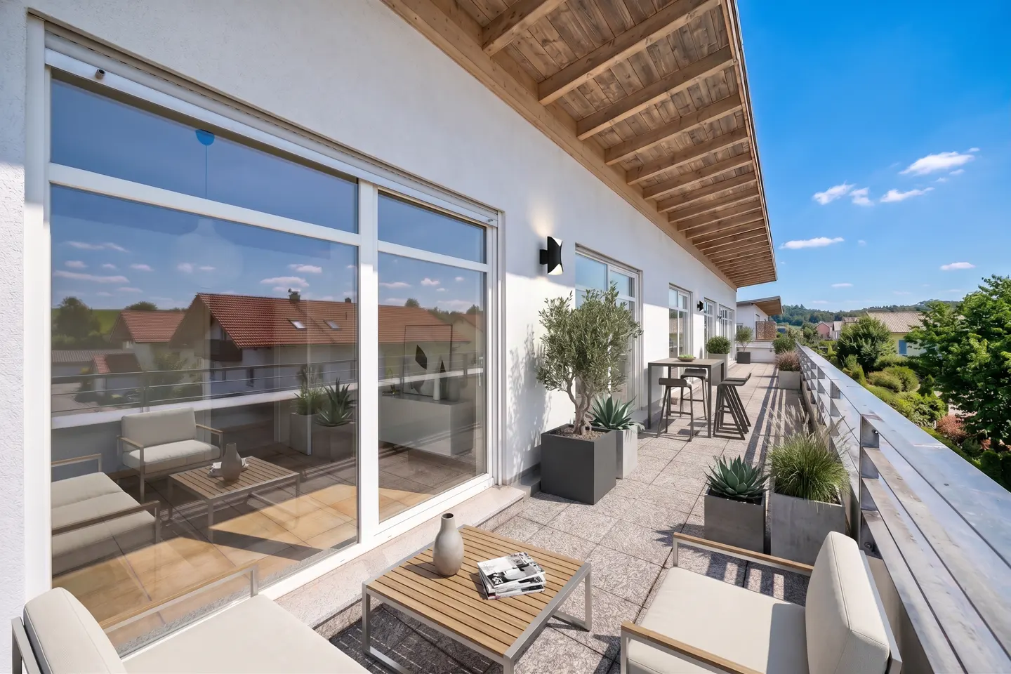 A long, sunny balcony with outdoor furniture, potted plants, and a view of trees and blue sky.