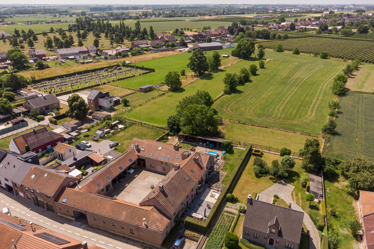 Aerial view of a U-shaped brick farmhouse with a central courtyard, surrounded by green fields and a rural village.