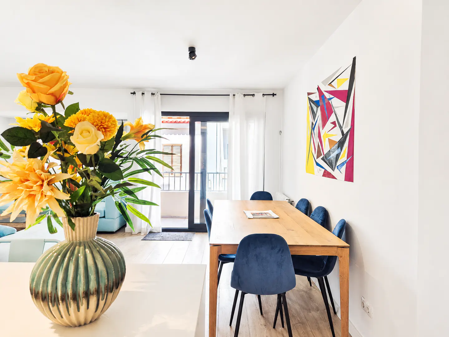 Bright dining room with a wooden table, blue velvet chairs, and a vase of yellow flowers. Abstract art hangs on the white wall. Balcony in background.