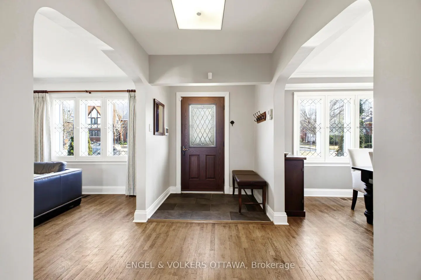 Foyer with hardwood floors, gray walls, and arched doorways. A dark wood door with a diamond-patterned window is centered. A bench sits to the right.