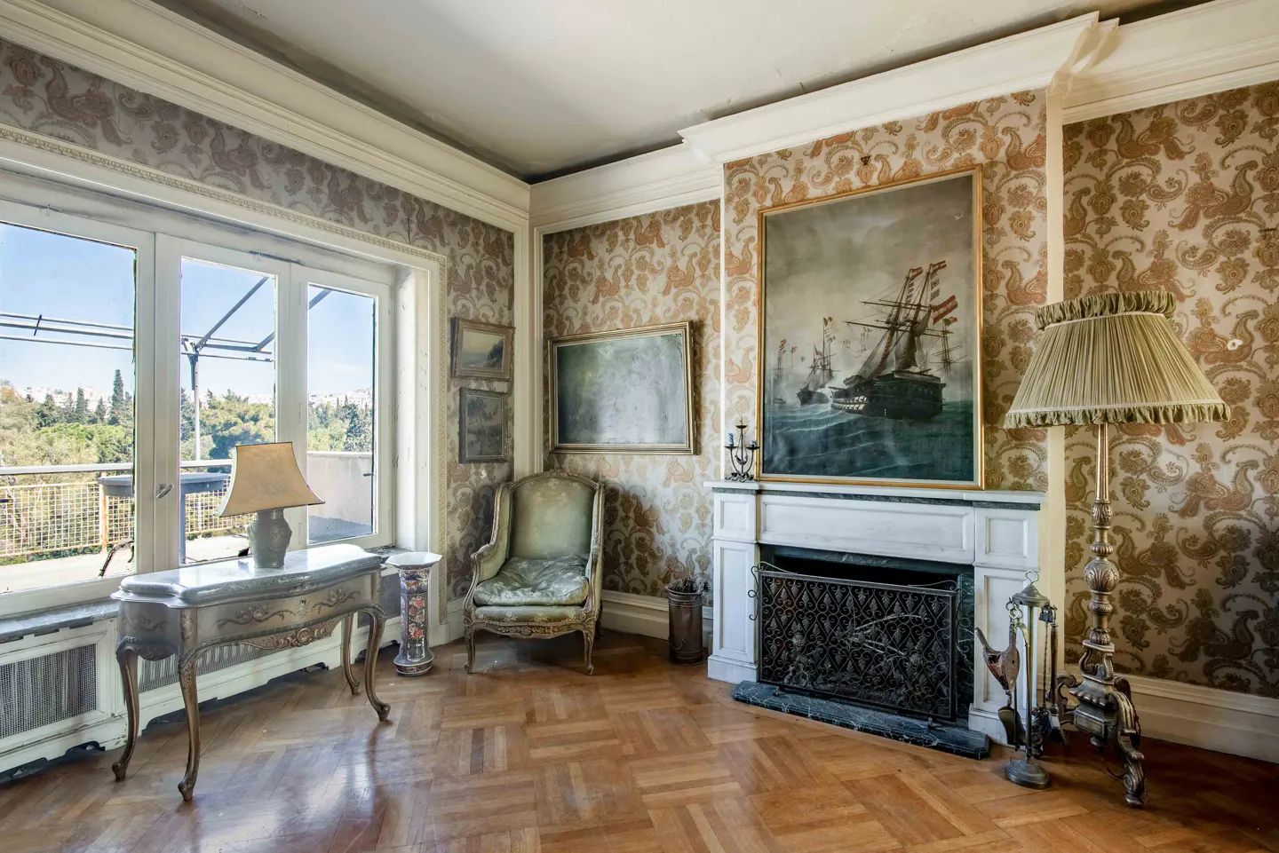 Ornate living room with patterned wallpaper, herringbone wood floors, and a large painting of ships over a marble fireplace.