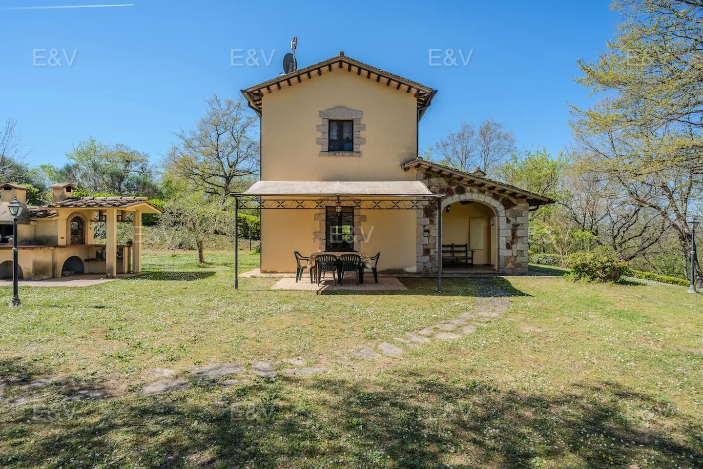 Exterior view of a two-story beige house with a stone archway and outdoor dining set on a green lawn.