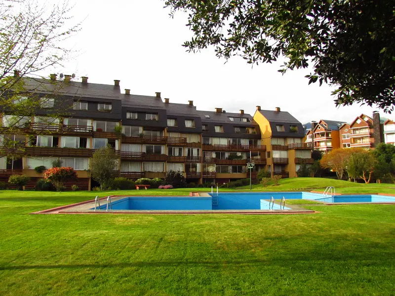 A multi-story apartment building with balconies overlooks two blue swimming pools on a green lawn.