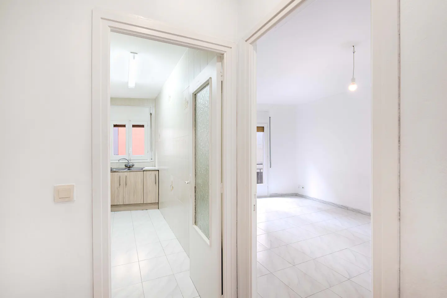 A bright, white apartment interior is seen through two doorways, revealing a kitchen and a living room with tiled floors.