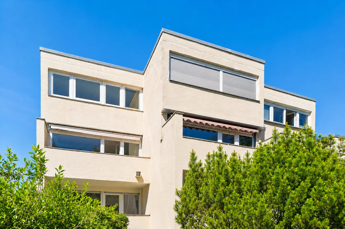 Modern beige apartment building with flat roof, large windows, and green trees against a clear blue sky.