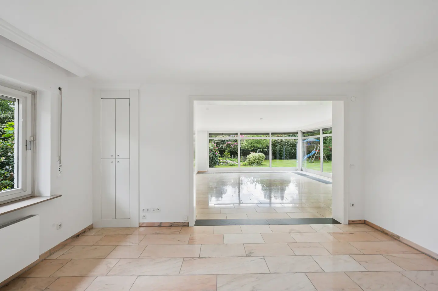 Bright, empty room with beige tile floor. A large opening leads to a sunroom and green garden with a blue slide. White walls and built-in cabinet.