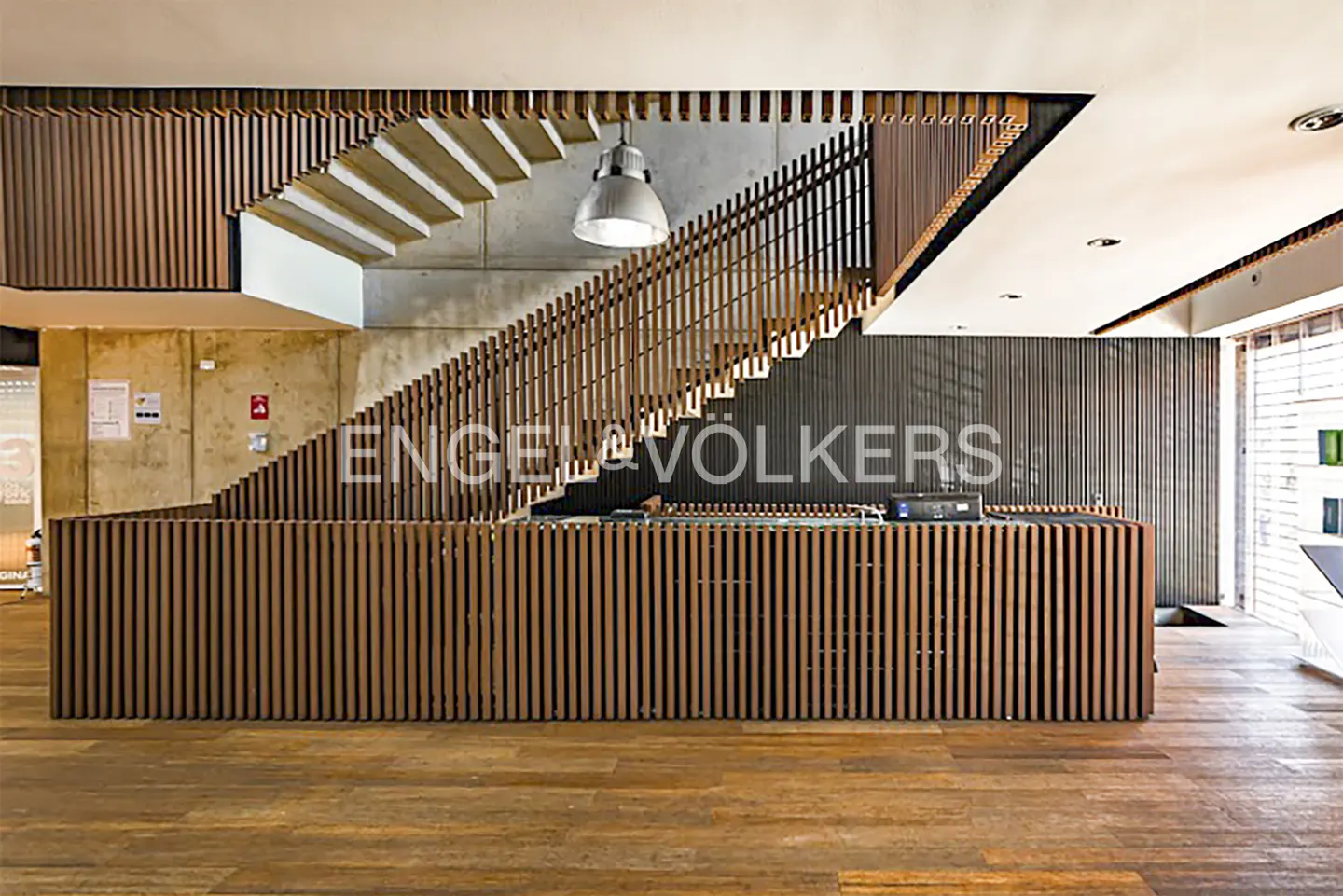 Engel & Völkers office interior with wood floors, a reception desk with vertical wood slats, and a staircase with wood railings.