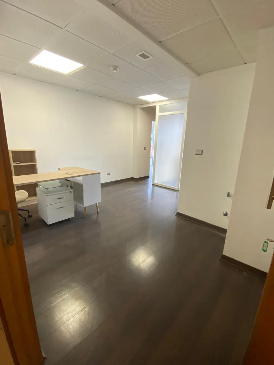 Bright, empty office space with dark wood floors, white walls, and a light wood desk with white drawers.