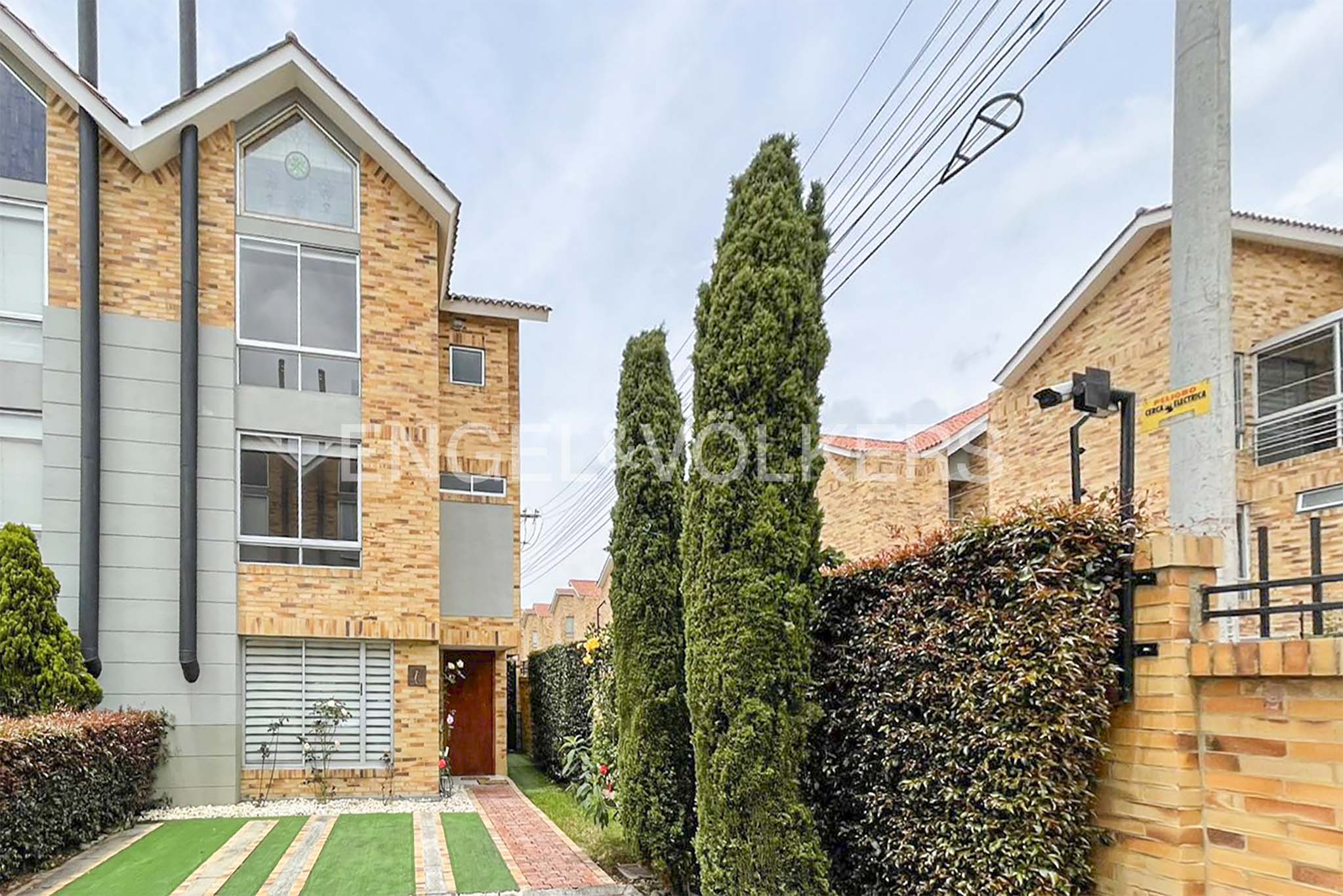 Exterior view of a brick townhouse with a brown door, green lawn, and tall trees. The sky is cloudy.