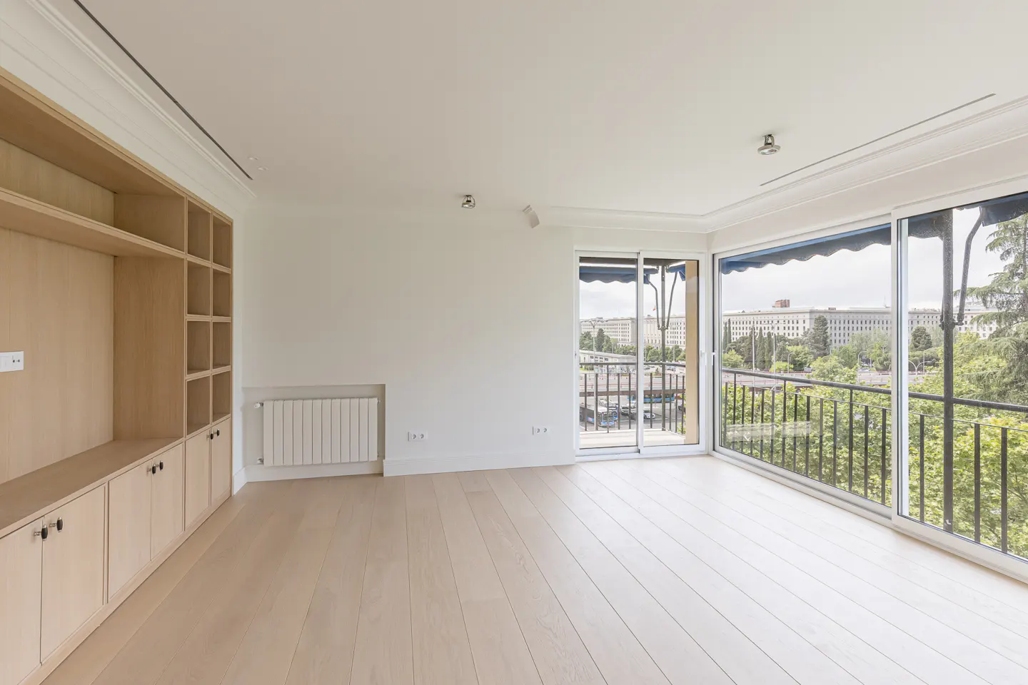 Bright, empty room with light wood floors, white walls, and a built-in bookcase. Balcony with city views through sliding glass doors.