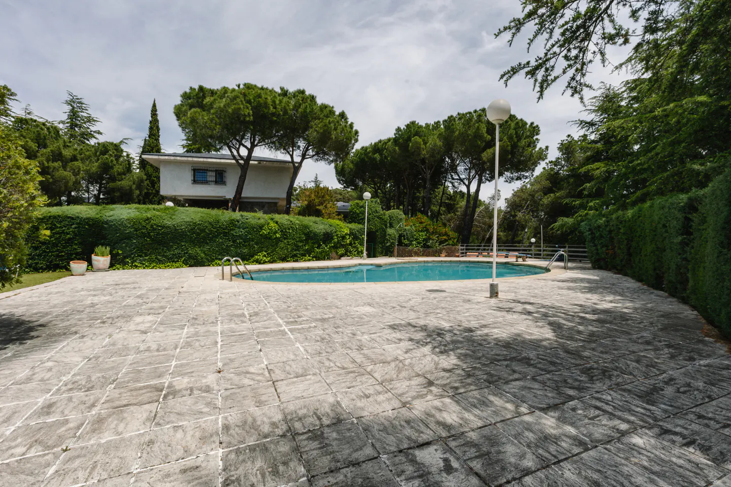 A backyard with a blue pool, a gray stone patio, green trees, and a white house in the background.