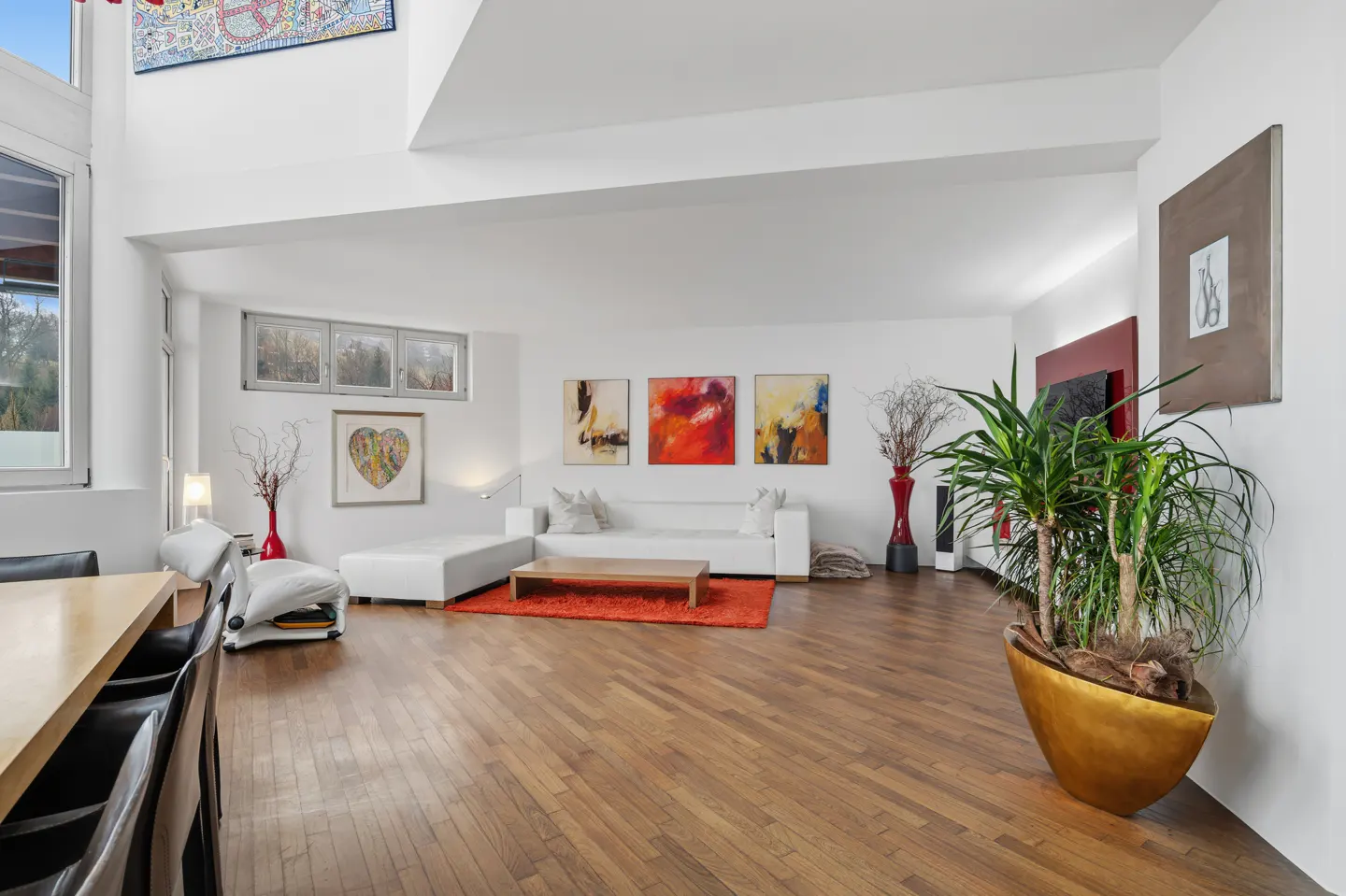 Bright, airy living room with hardwood floors, white walls, and modern art. A white sectional sofa sits on an orange rug. A large potted plant adds a touch of green.