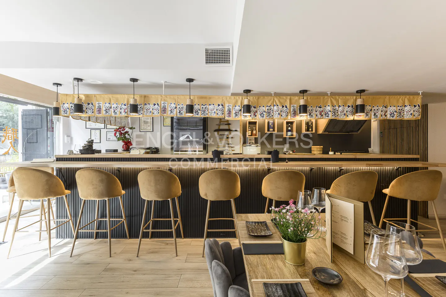 Interior of a restaurant with a bar, tan stools, and wooden tables set with glassware and menus. Overhead lighting and a decorative valance.