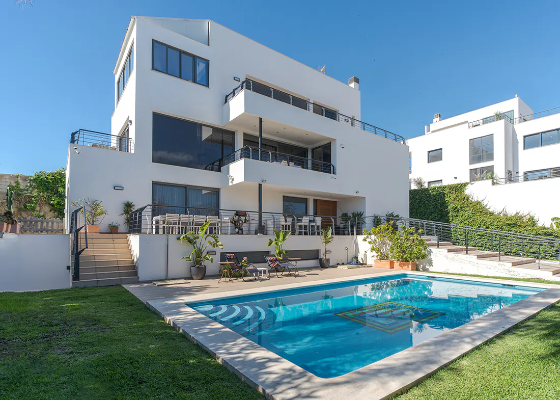 Back view of a modern, white, multi-story house with a pool and green lawn on a sunny day.