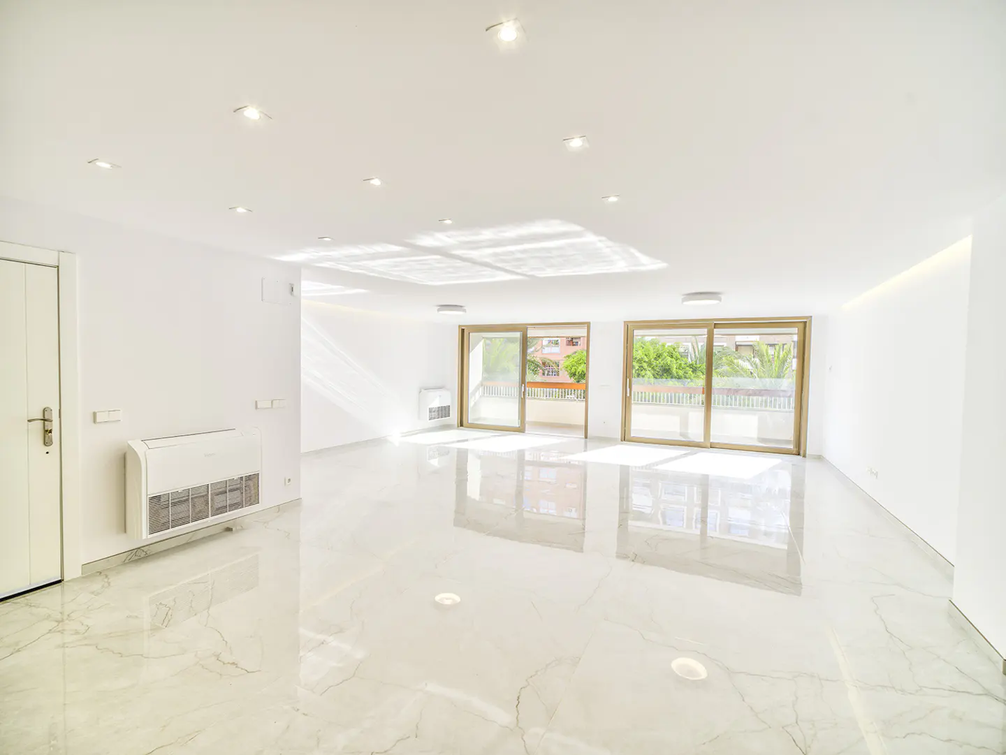 Bright, empty room with white walls, marble floor, and recessed lighting. Two sliding glass doors lead to a balcony with green trees visible.