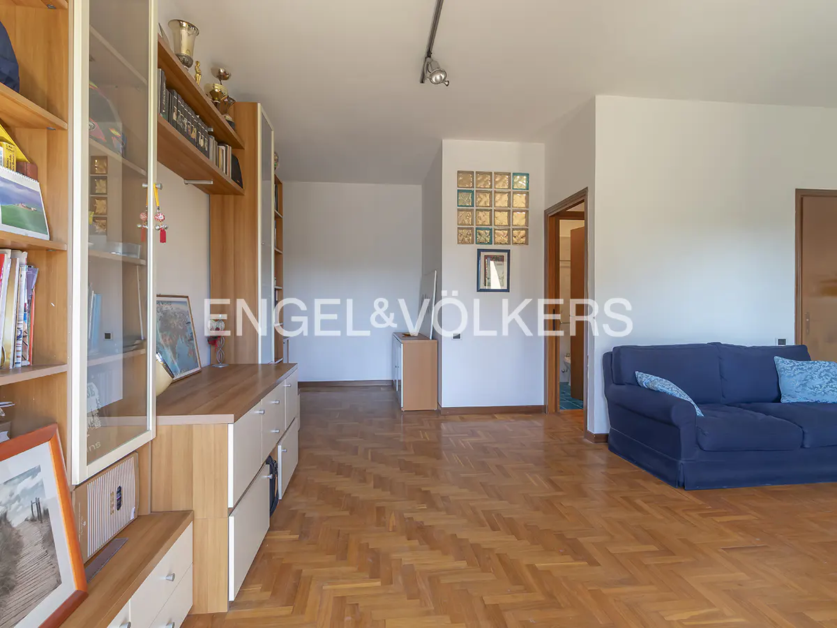A bright living room with parquet floors, a blue sofa, and a built-in wooden bookshelf. The Engel & Völkers logo is visible.