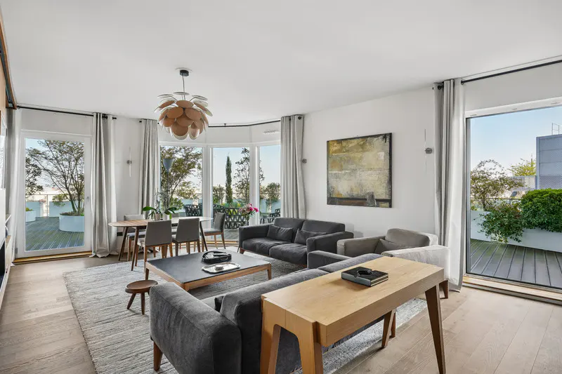 Bright living room with gray sofas, wood floors, and a large rug. Windows open to a terrace with greenery. Modern light fixture and abstract art.