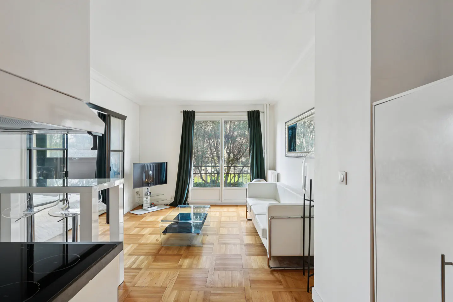 Modern living room with white sofa, glass coffee table, parquet floors, and balcony doors with dark green curtains.
