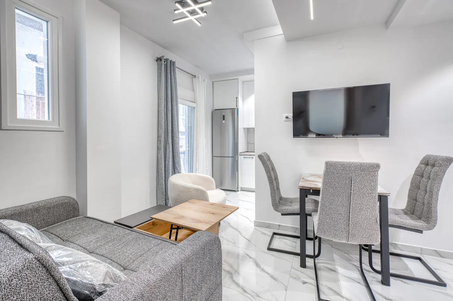 Bright, modern living room with gray sofa, dining table with chairs, and a flat-screen TV on a white wall. A window and a white armchair are also visible.