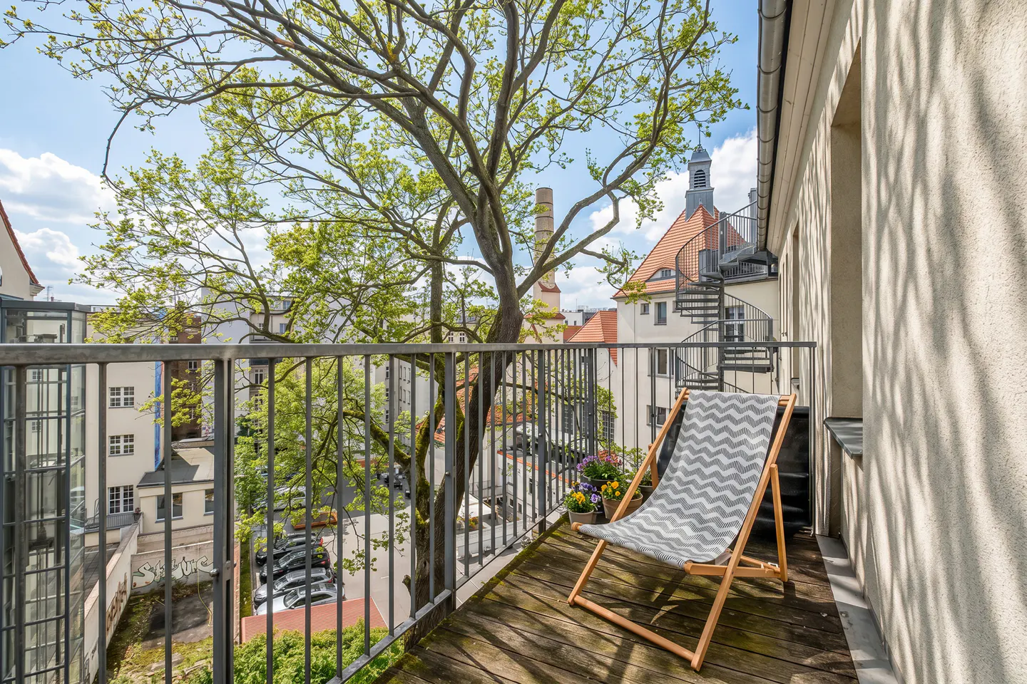Balcony view with a gray chevron lounge chair, potted flowers, and a metal railing overlooking a tree and buildings.