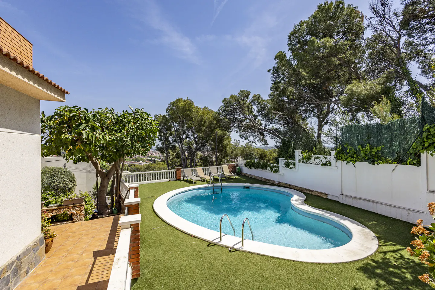 Outdoor kidney-shaped pool with turquoise water, surrounded by green turf, trees, and a white fence under a blue sky.