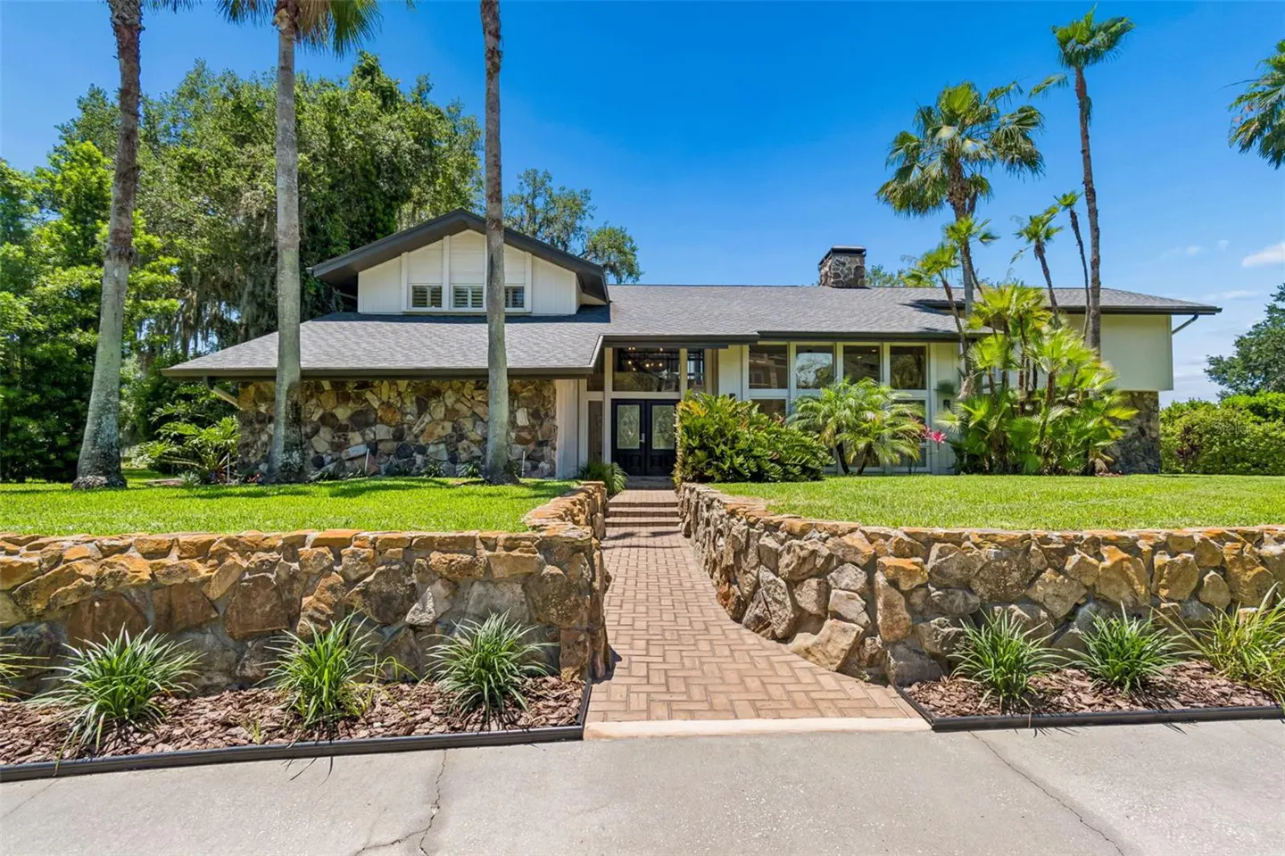 Exterior of a large, modern home with stone accents, a brick walkway, and palm trees under a blue sky.