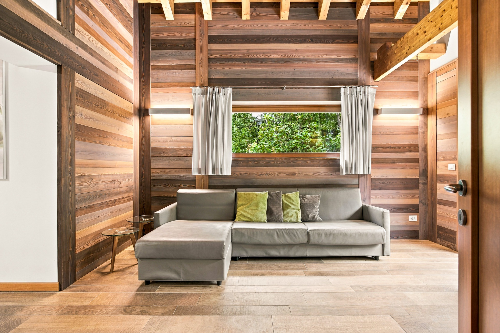 Modern living room with wood-paneled walls, a grey sectional sofa with green pillows, and a window overlooking greenery. [AI-generated text]