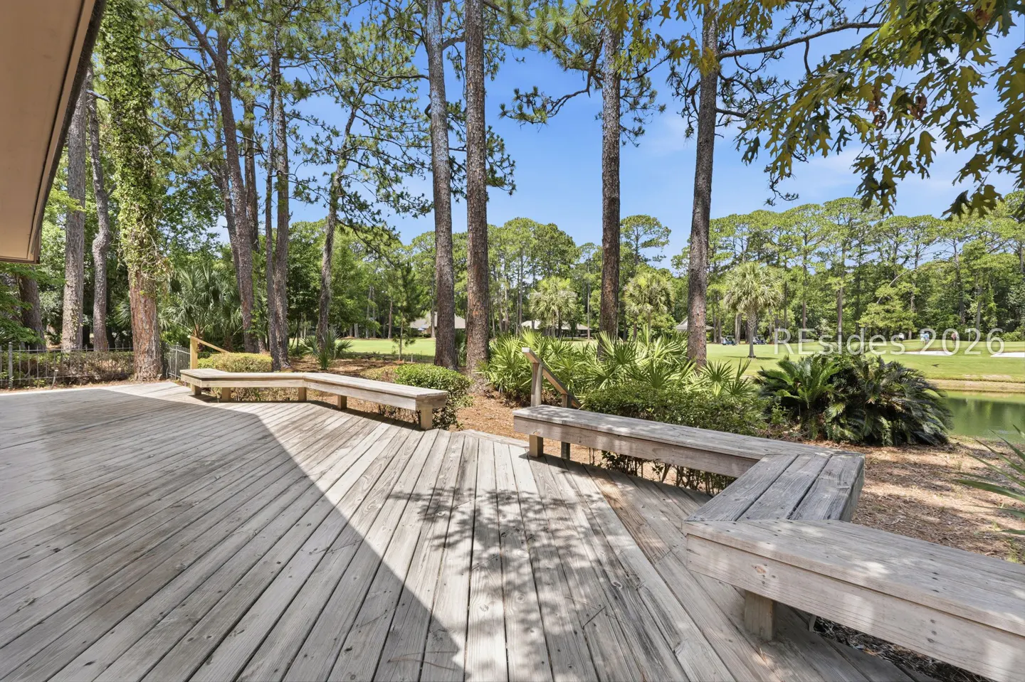 Wooden deck with built-in benches overlooking a pond, lush green trees, and a golf course under a clear blue sky.