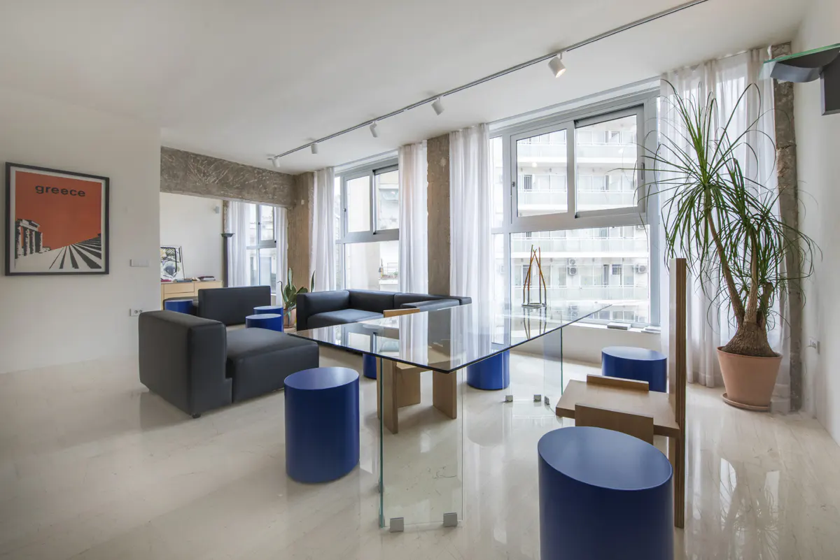 Bright living room with gray sofas, blue stools, and a glass table. Large windows with white curtains and a potted plant add natural light.