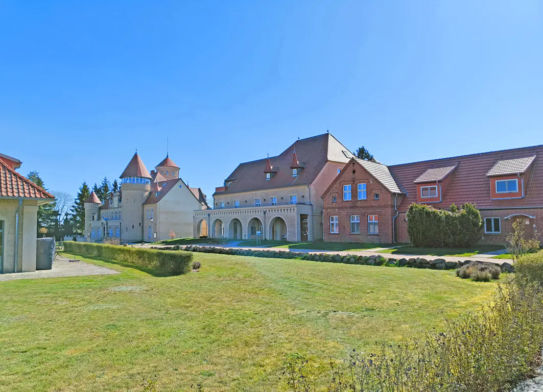 Exterior view of a castle-like building with red roofs, turrets, and arched walkways, surrounded by a green lawn under a clear blue sky.