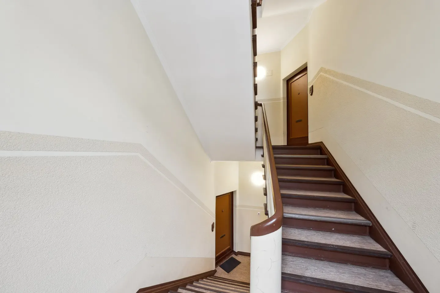 Interior view of a stairwell with brown wooden stairs and a white banister. Cream-colored walls and brown doors are visible on each landing.