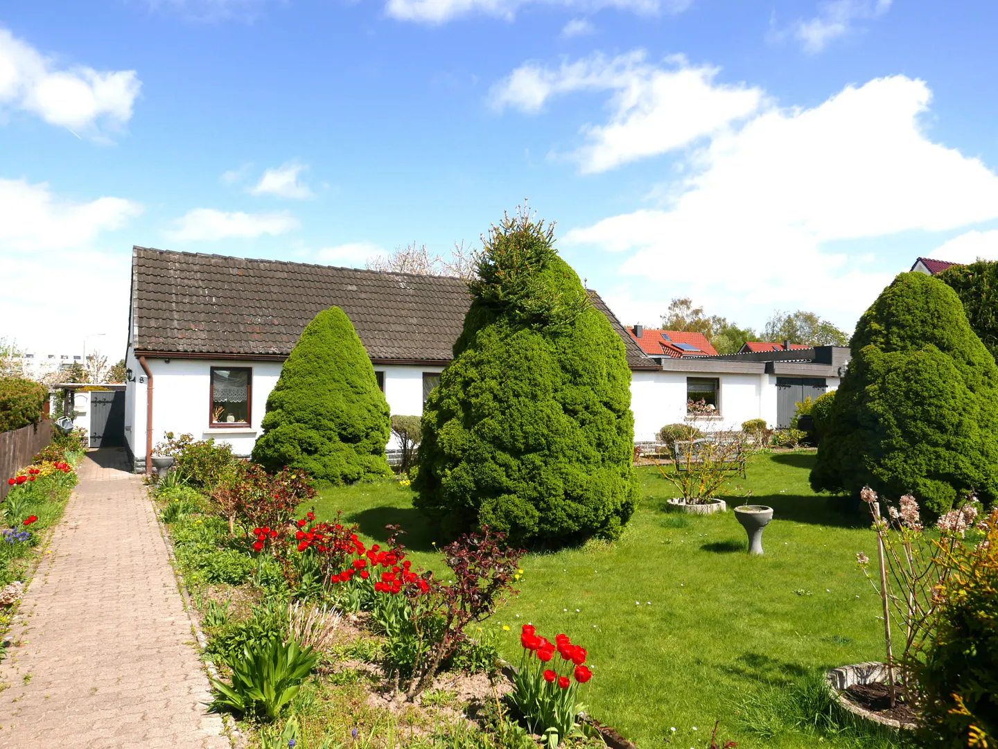 A white house with a gray roof is surrounded by green trees, grass, and red flowers under a blue sky.