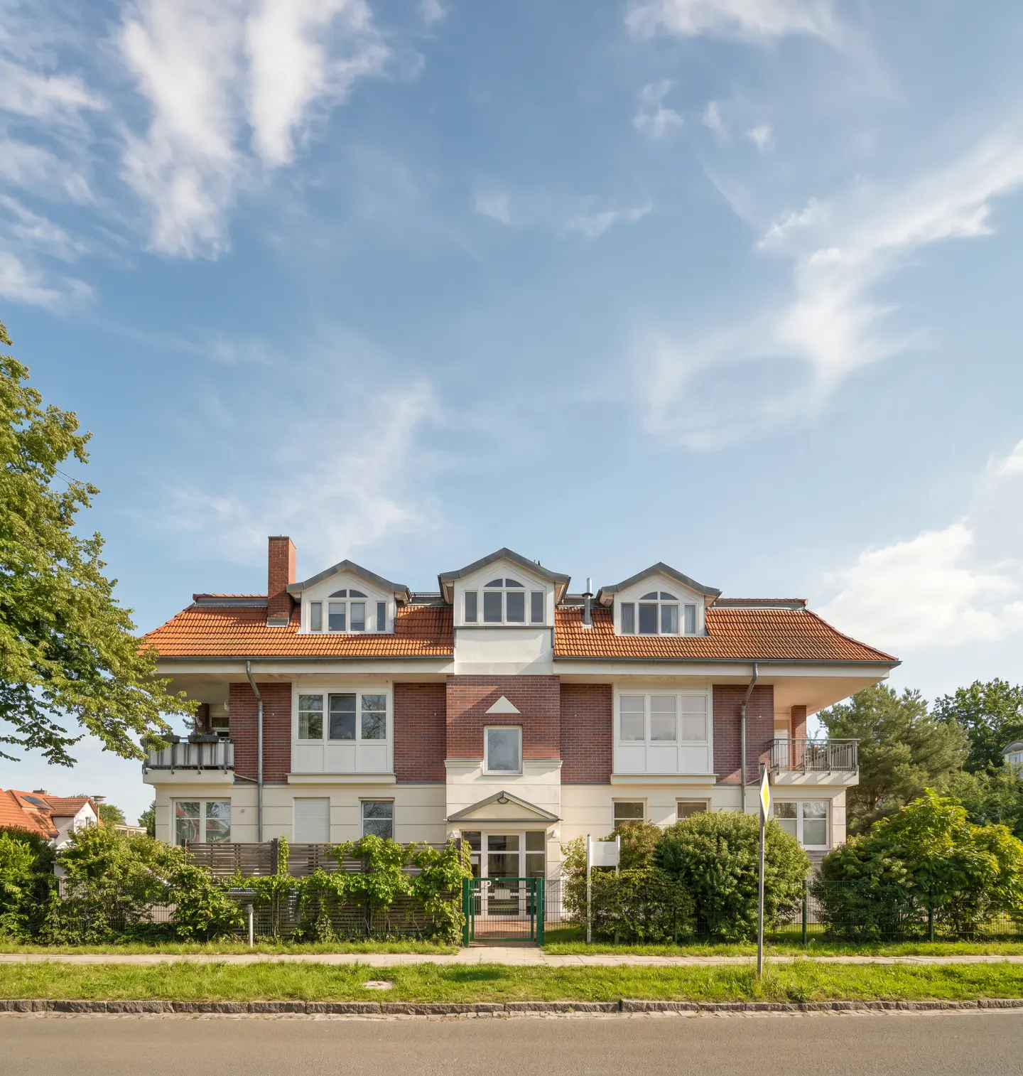 Two-story brick and stucco building with a red tile roof under a blue sky. Green bushes line the front of the property.
