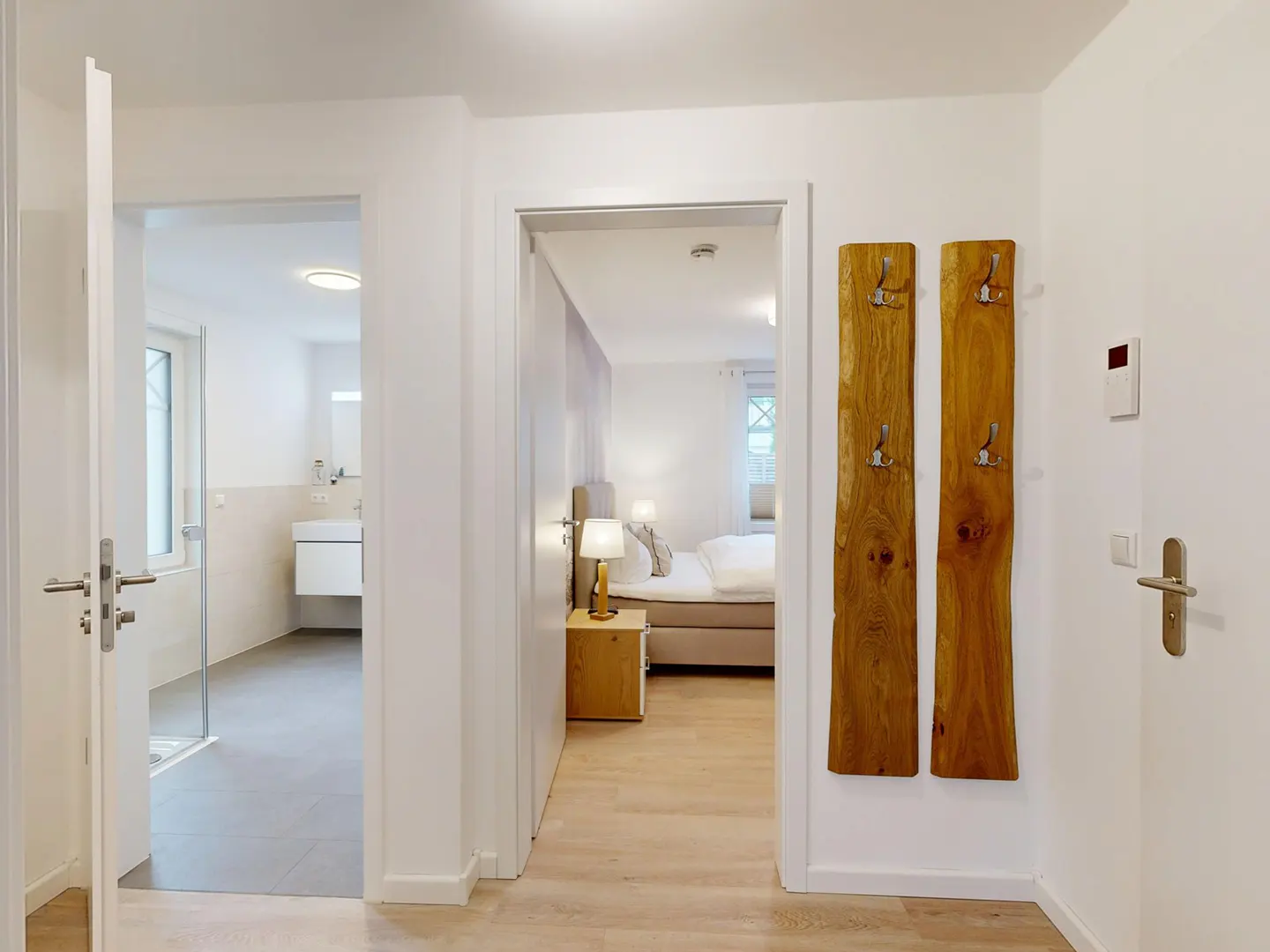 Hallway with open doorways to a bathroom and bedroom. Two wooden coat racks hang on the white wall.